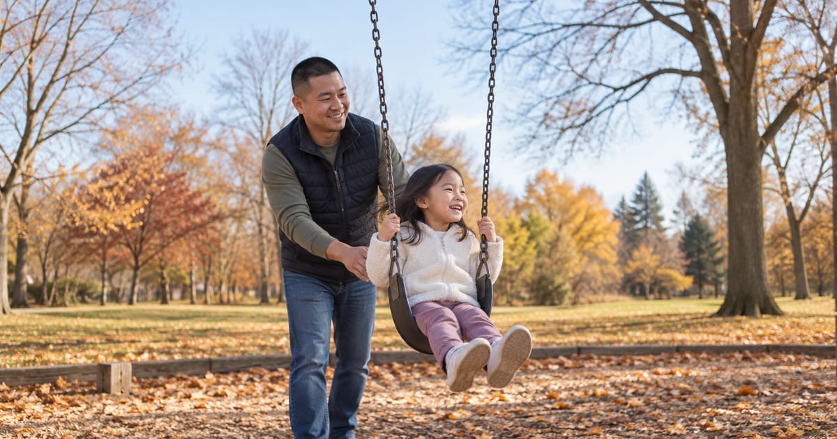 A smiling man in a quilted vest pushes a laughing young girl on a swing in a park strewn with fallen autumn leaves